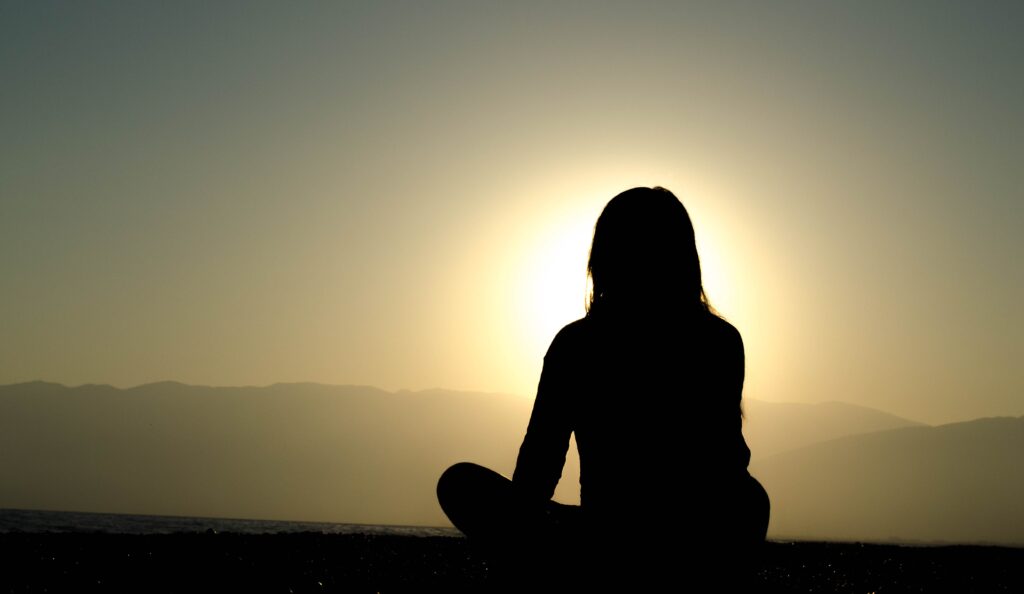 A person sitting cross-legged in meditation at sunrise with mountains and a calm sea in the background, symbolizing mindfulness and serenity.