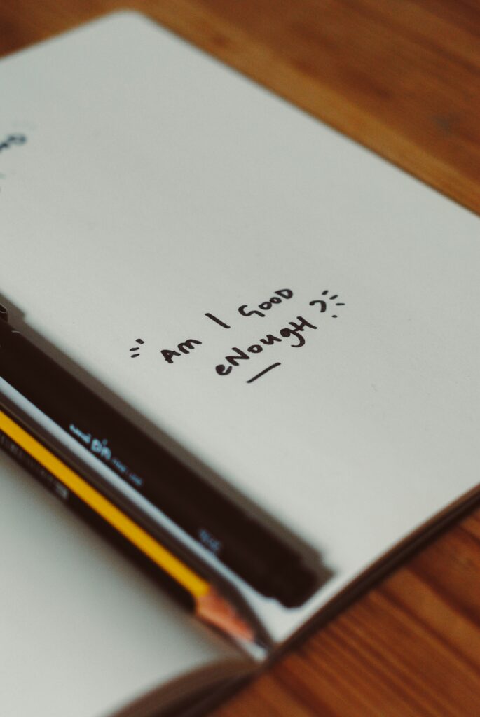 A notebook page with the handwritten question 'Am I good enough?' alongside a pencil and pen on a wooden desk.