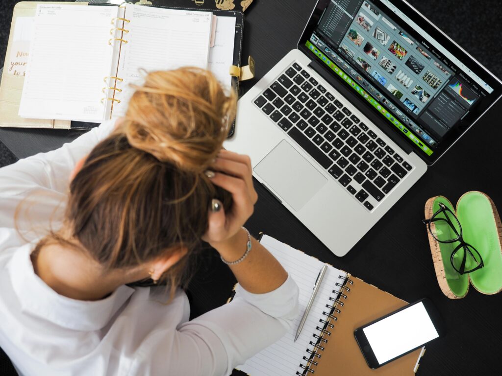 A stressed woman sitting at a desk with her head in her hands, surrounded by a laptop, planner, notebook, and phone, symbolizing perfectionism and overwhelm.