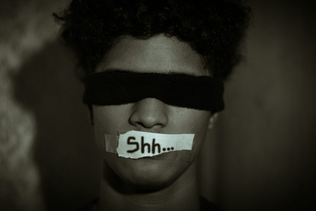 Person with a blindfold over their eyes and tape over their mouth labeled "Shh...", illustrating the shame and silencing around mental health struggles.