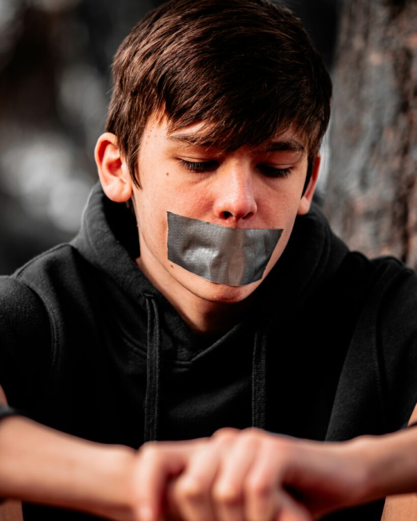 Teenage boy with duct tape over his mouth, sitting silently with a downcast gaze, representing the silencing effects of mental health stigma.