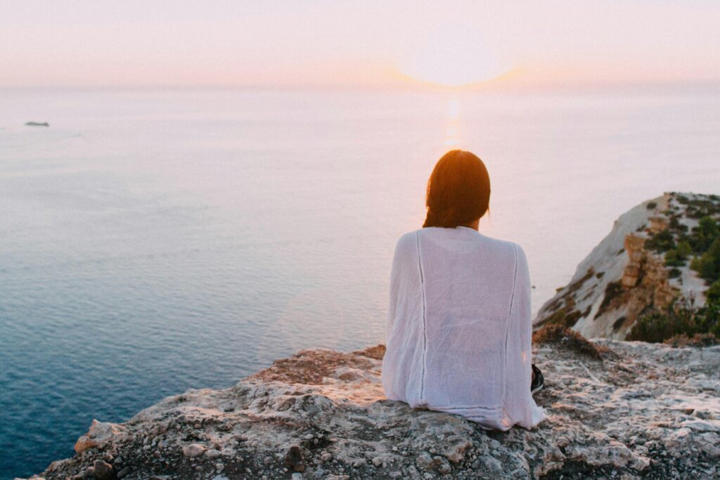 Woman sitting on a cliff at sunset, looking out over the ocean—representing calm, reflection, and caring for mental health before reaching a crisis point.