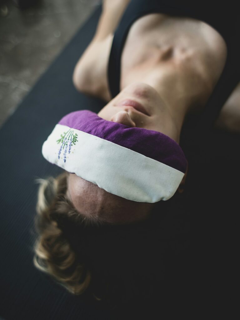 Woman lying on a yoga mat with a lavender-filled eye pillow across her eyes, practicing deep rest and nervous system regulation.
