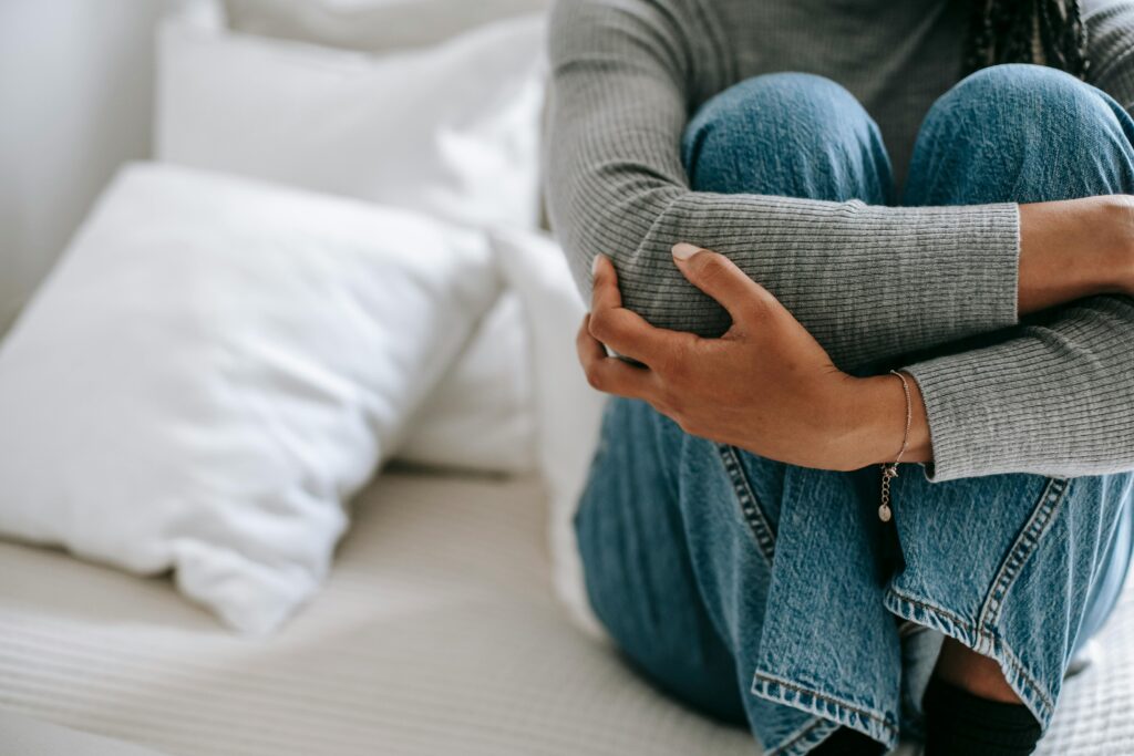 Body of a woman sitting on a bed curled up holding her legs in a dorsal vagal nervous system state.